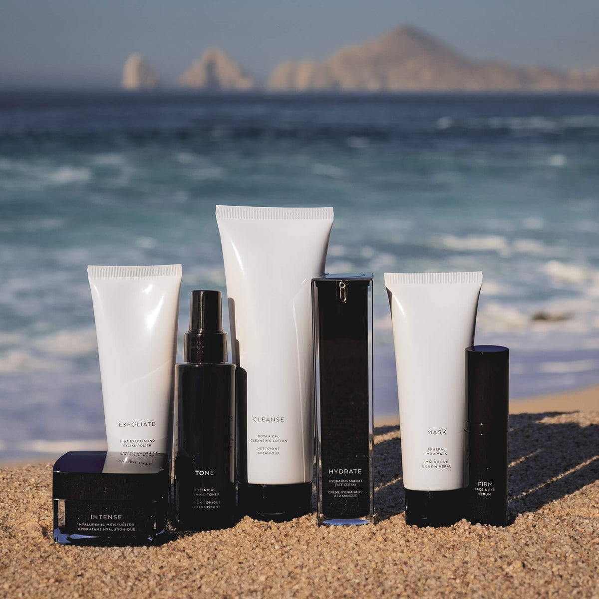 The Barrier Balm displayed with a full lineup of matching skincare bottles and tubes on a beach with ocean and cliffs in the background